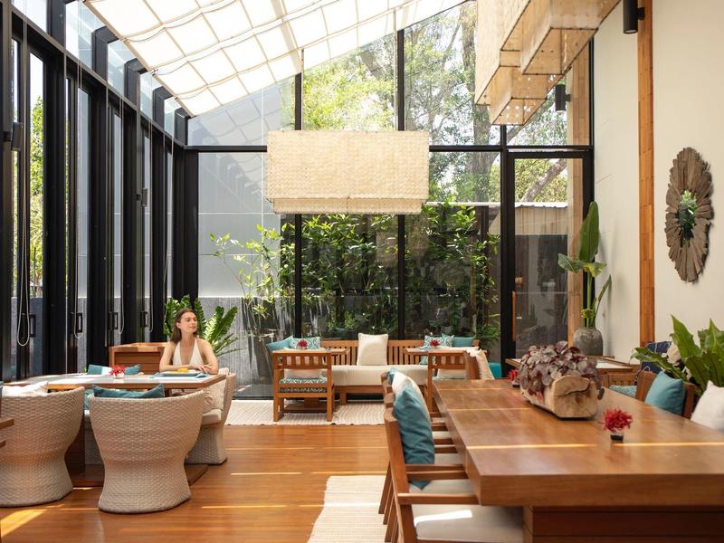 Lichte eetkamer met glazen wand, houten tafels en stoelen, decoratieve planten en een persoon.