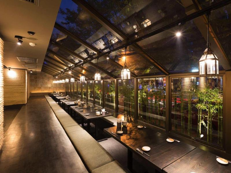 Langes, modernes Restaurant mit Glasdach, beleuchteten Tischen und gemütlichen Sitzbänken bei Nacht.