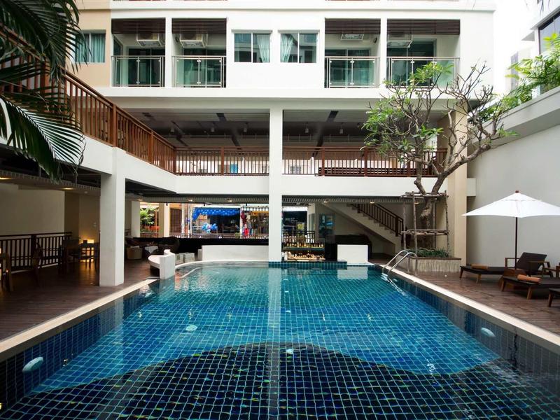 Moderner Hotelpool mit blauen Fliesen, Liegestühlen, Sonnenschirmen und umliegenden Gebäuden.