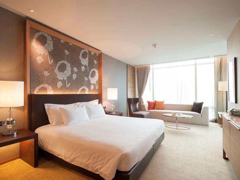 Helles modernes Schlafzimmer mit großem Bett, floralem Kopfteil und Sofa vor großen Fenstern.