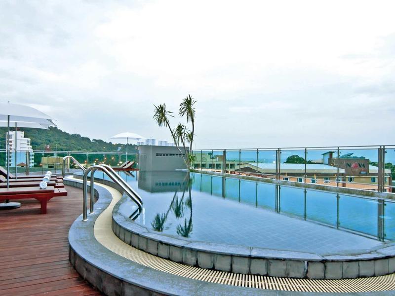 Runder Infinity-Pool mit Holzdeck, Liegestühlen und palmengesäumter Meereskulisse bei bewölktem Himmel.