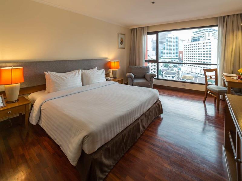 Modern hotel room with large double bed, wooden floor, and city view through large window.