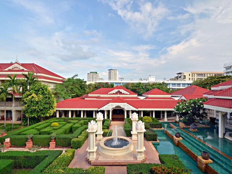 Hotel garden with fountain, red-roofed buildings, and manicured green hedges beneath a bright sky.