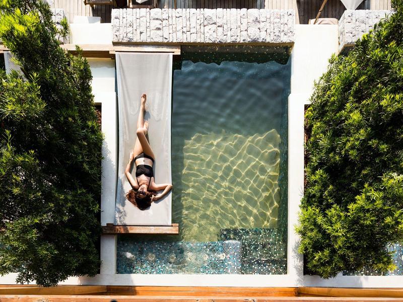 Eine Person liegt auf einer Liege neben einem klaren rechteckigen Schwimmbecken, umgeben von Bäumen.