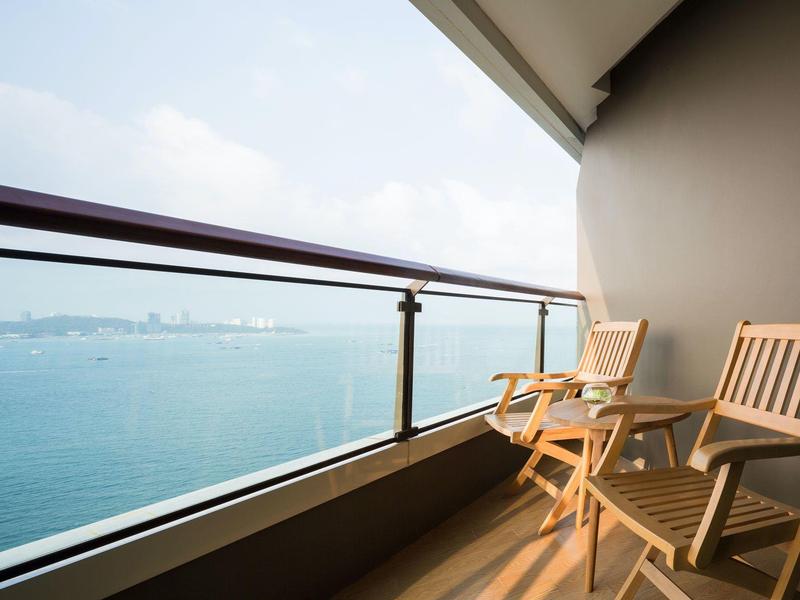 Balkon mit zwei Holzstühlen und Glasgeländer, Blick auf ruhiges, blaues Meer unter hellblauem Himmel.