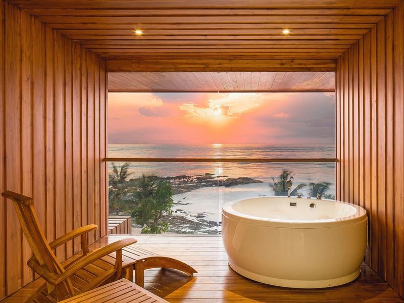 Holzterrasse mit Rundbadewanne und zwei Stühlen, Blick auf Strand und Sonnenuntergang.