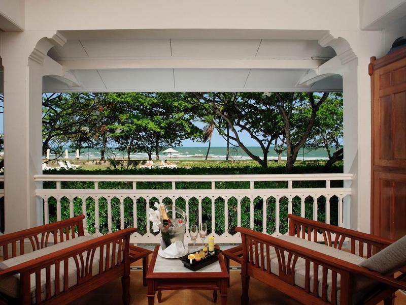 Balkon met houten stoelen, tafel en uitzicht op de oceaan geframed door bomen en wit hek.