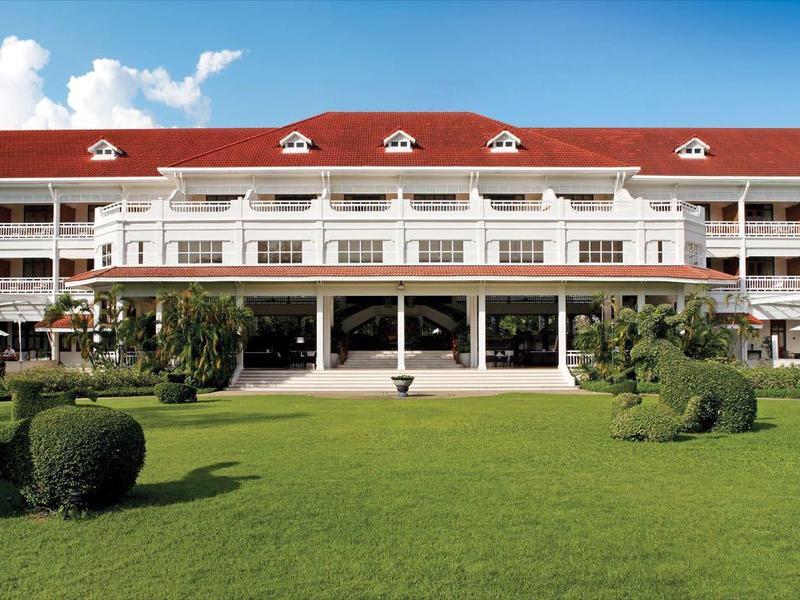 Een groot hotel met een gebogen gevel, rood dak, weelderig groen gazon en gesnoeide struiken onder een blauwe lucht.