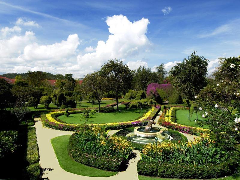 Heldere tuin met groene struiken, kronkelpaden en een centrale fontein onder een bewolkte lucht.