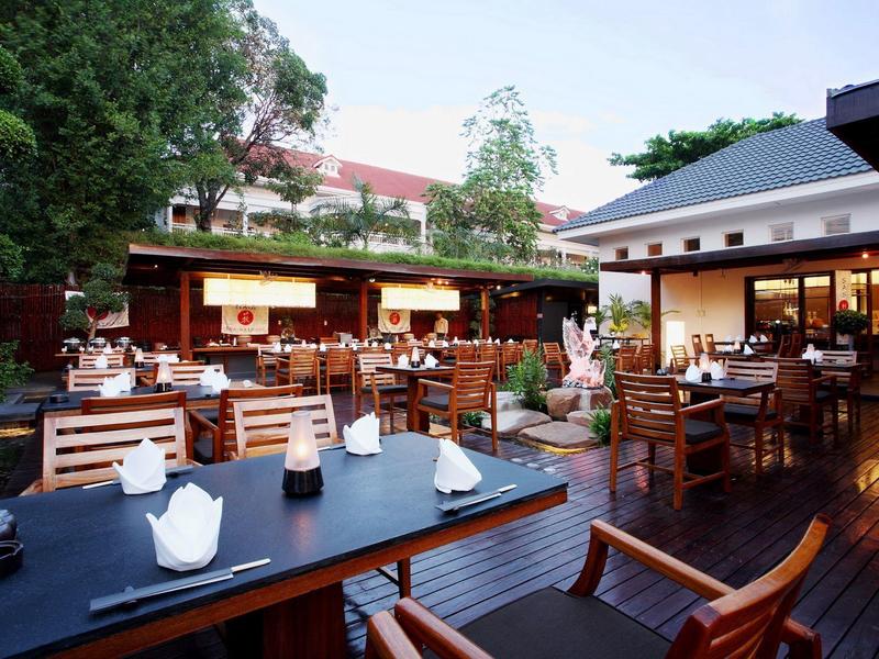 Buiten eetgedeelte met houten tafels en stoelen omgeven door groen en gebouwen.