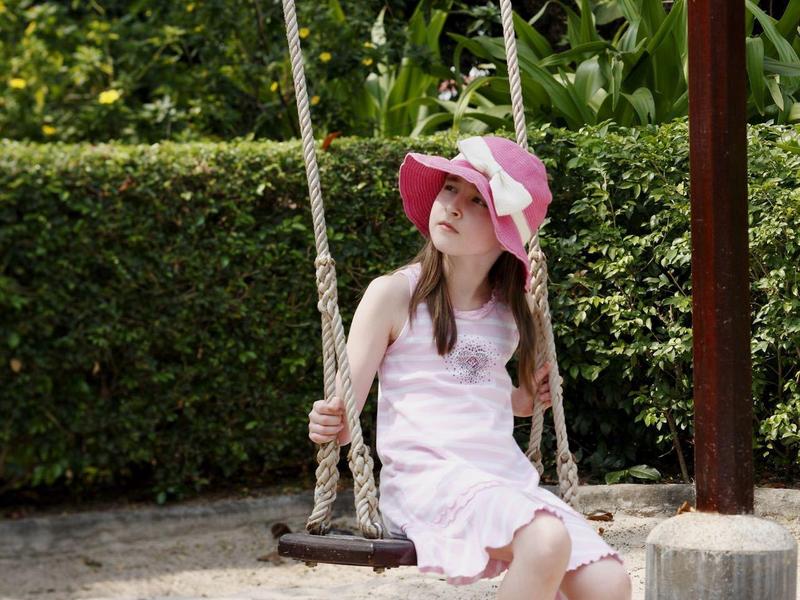 Meisje in witte jurk en roze hoed zit blootsvoets op een houten schommel in een tuin.