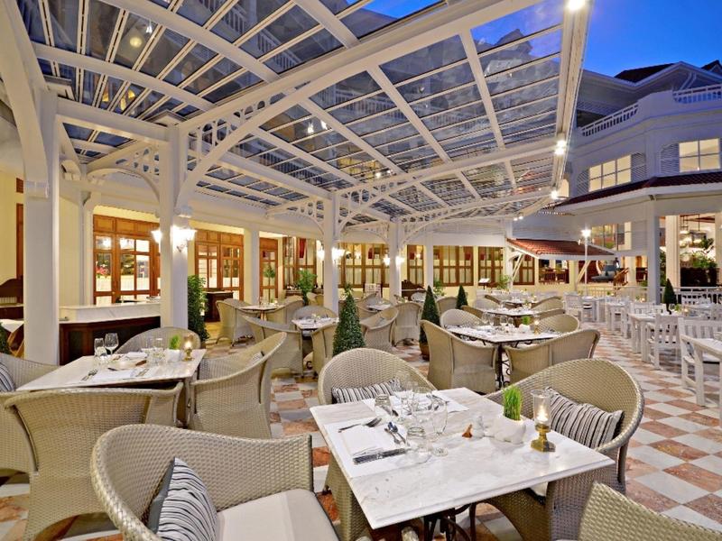 Elegant buitenrestaurant met witte tafels, beige stoelen en glazen dak onder avondhemel.