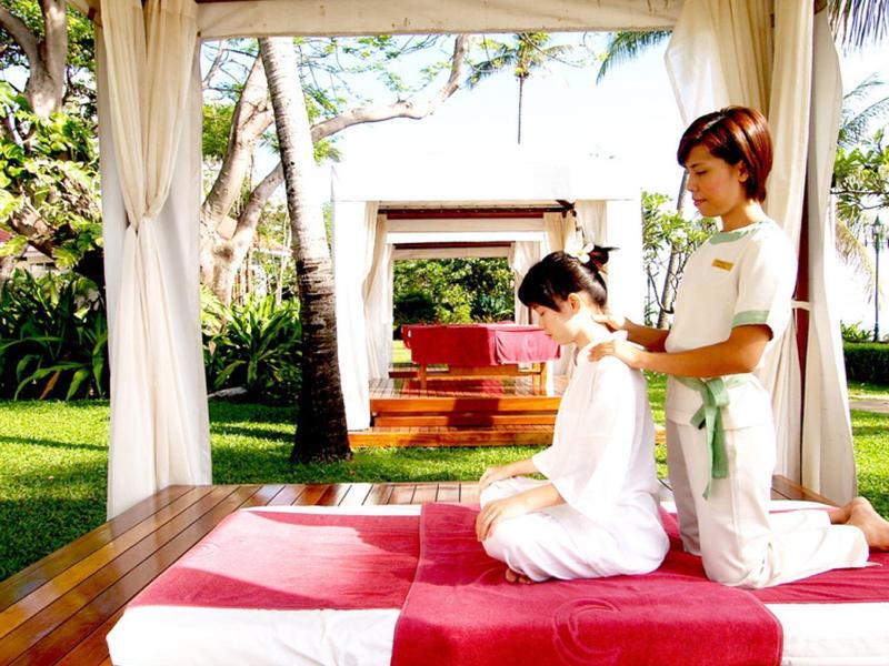 Twee personen genieten van een zittende massage onder een houten overkapping met kussens en groen.