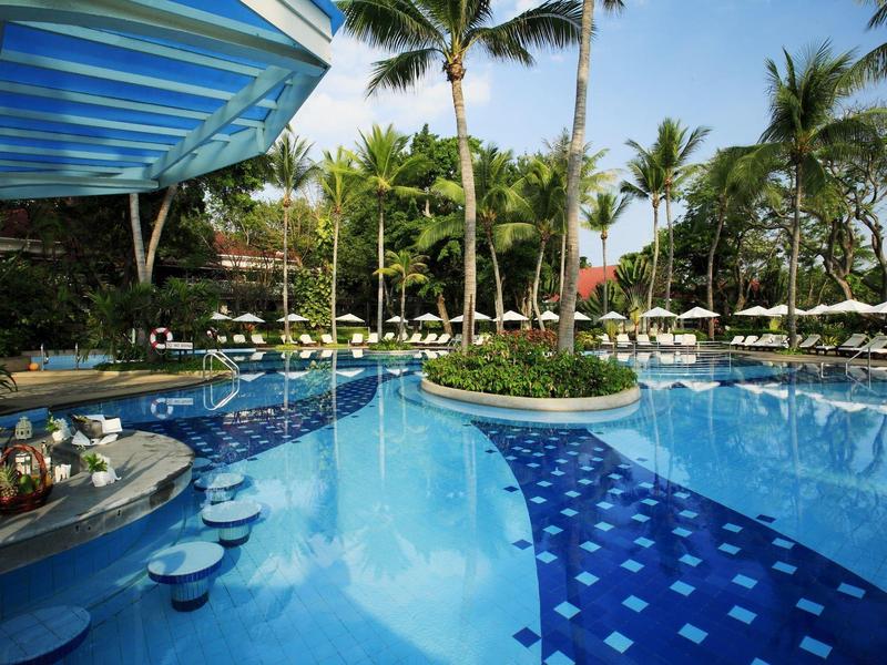 Buitenzwembad met palmbomen en blauwe lucht, met krukjes in het water en ligstoelen eromheen.