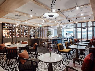 Modern eingerichteter Gastraum mit Holzdecke, bunten Sesseln und geometrischem Teppich.