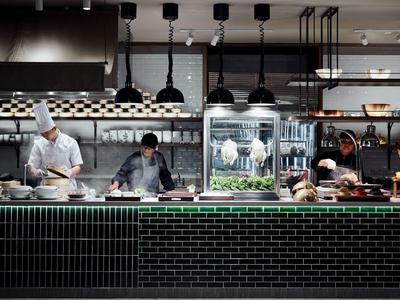 Drei Köche bereiten in moderner Küche mit schwarzen Fliesen und Lampen Essen zu.