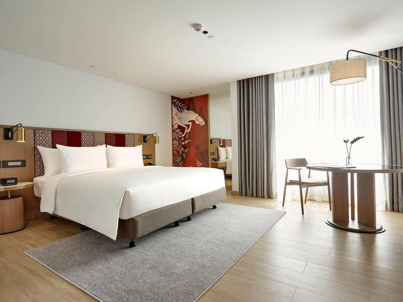 Modernes Hotelzimmer mit großem Bett, Teppich, Tisch, Stühlen und bodentiefen Fenstern mit Vorhängen.