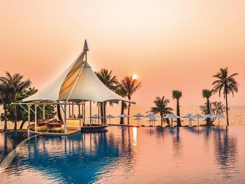 Infinity-Pool mit weißem Pavillon und Palmen, Sonnenuntergang im Hintergrund, ruhiges Wasser reflektiert Licht.