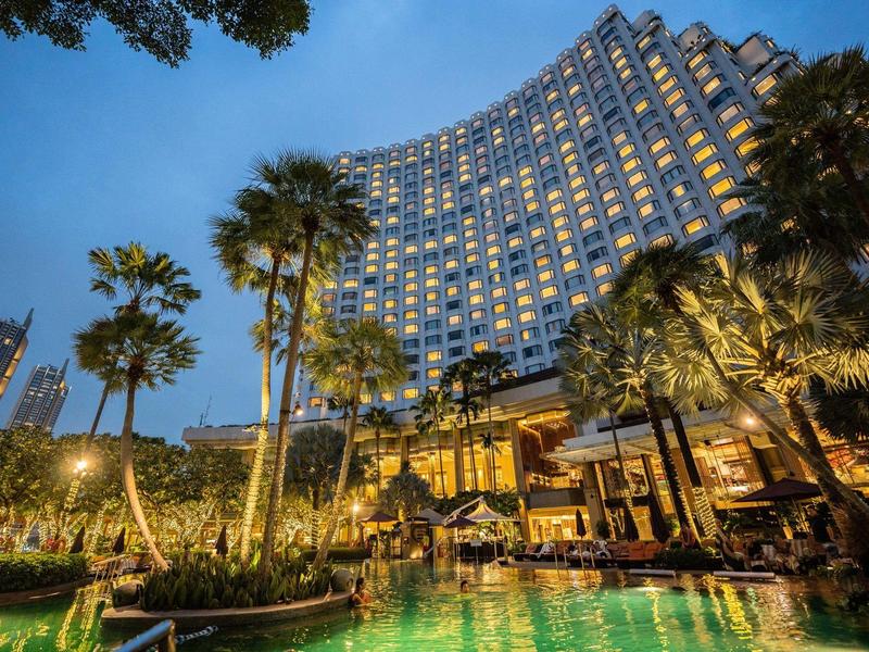 Hochhaus mit vielen beleuchteten Fenstern, umgeben von Palmen und beleuchtetem Pool bei Abenddämmerung.