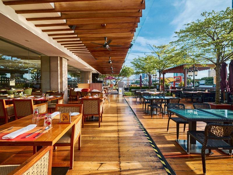 Offenes Restaurant mit Holzterrasse, Tischen, Stühlen und Bäumen bei sonnigem Himmel.