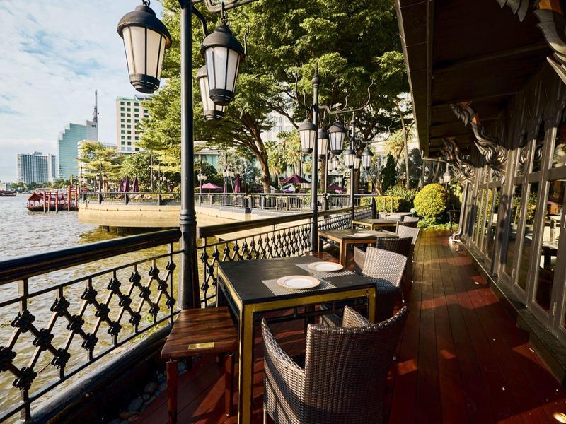 Einladende Terrasse mit Tischen und Stühlen am Flussufer, umgeben von Bäumen und Gebäuden.