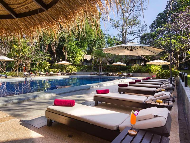 Poolbereich mit Liegen, Sonnenschirmen und tropischer Bepflanzung an einem sonnigen Tag.