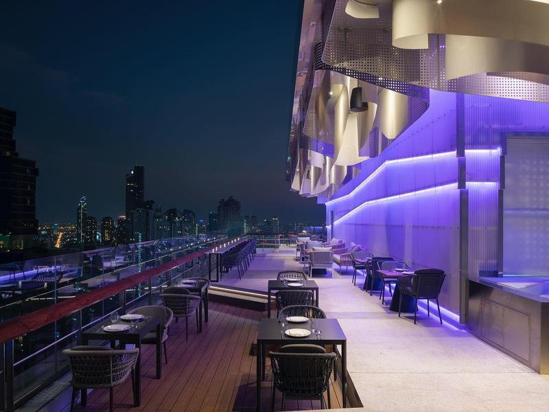 Dachterrasse mit Tischen, Stühlen und beleuchteten violetten Fassadenelementen bei Nacht.