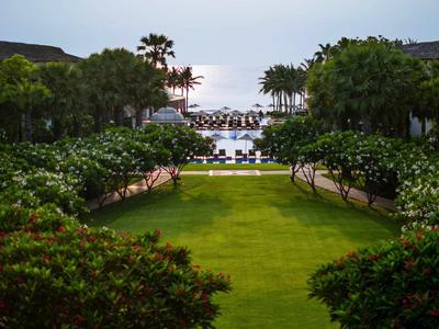 Blick auf einen gepflegten Garten zu einem Strand mit Palmen und Lounge-Bereichen am Wasser.