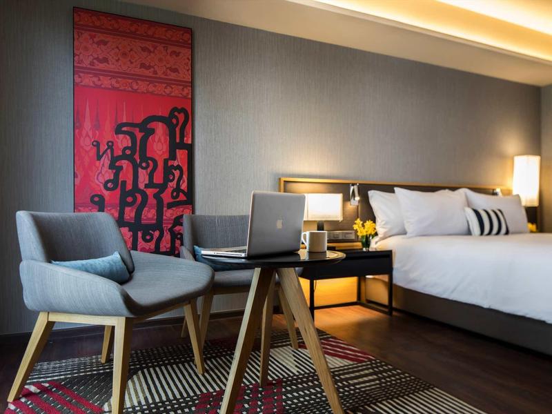 Modernes Schlafzimmer mit großem Bett, blauem Sessel, Tisch, Laptop und rotem Kunstbild an der Wand.