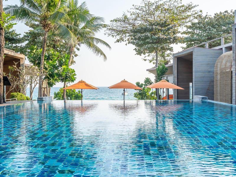 Infinity-Pool mit blauem Wasser, Palmen, Sonnenschirmen und Meer im Hintergrund bei klarem Himmel.