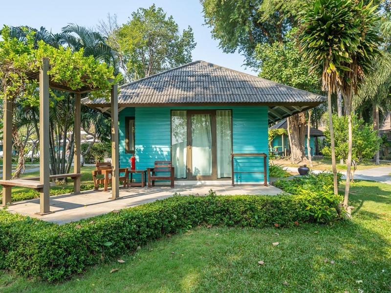 Kleines türkisfarbenes Haus mit Terrasse, umgeben von Grün, Büschen und Bäumen bei Sonnenschein.