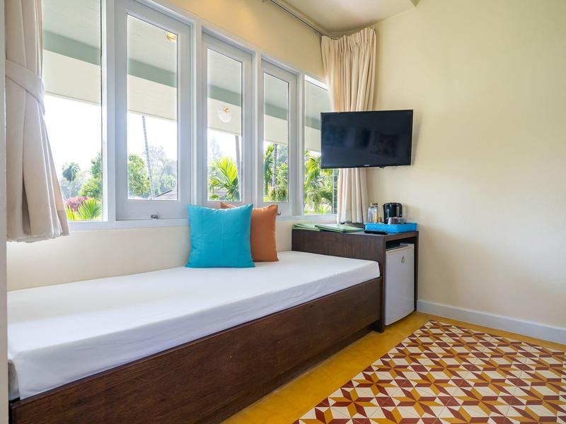 Heller Raum mit dunkelbraunem Tagesbett, bunten Kissen, Fernsehgerät, Minikühlschrank und gemustertem Teppich.