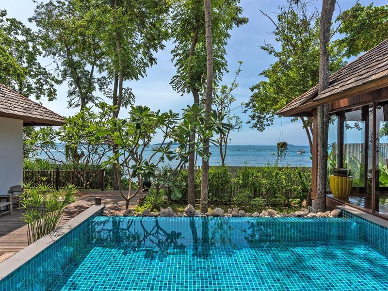 Blick auf einen blauen Pool mit Bäumen und Meer im Hintergrund bei klarem Himmel.