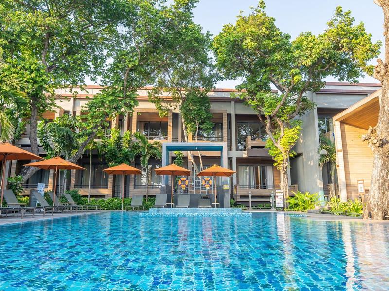 Hotelpool mit Liegestühlen und Sonnenschirmen, umgeben von grünen Bäumen und modernem Gebäude.