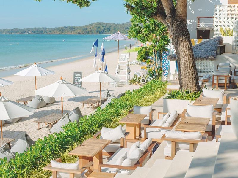 Strand mit weißen Sonnenschirmen, Holzliegen und Tischen, dahinter ruhiges blaues Meer und Himmel.