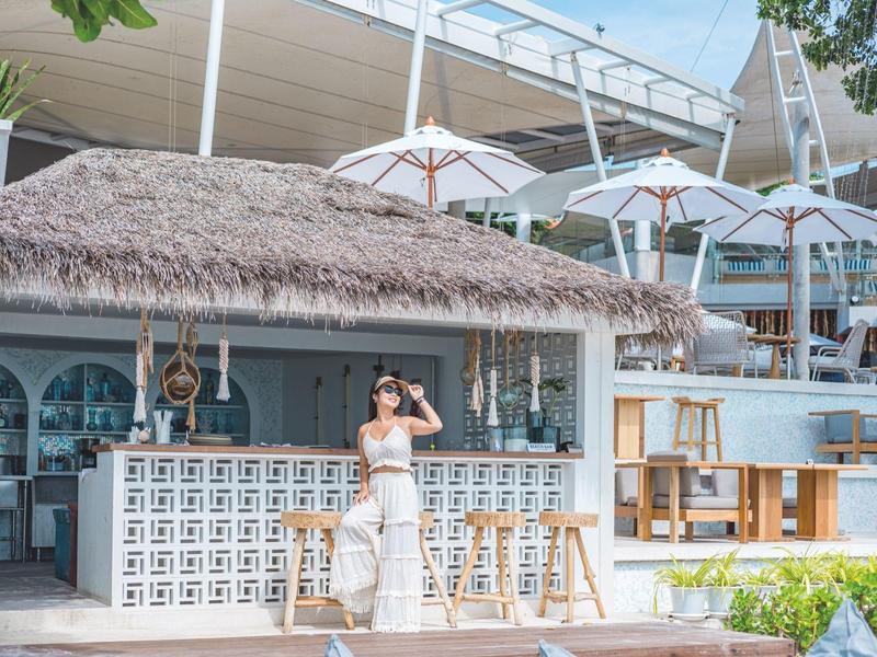 Strandbar mit Strohdach, weißen Barhockern und Sonnenschirmen, Frau lehnt entspannt am Tresen.