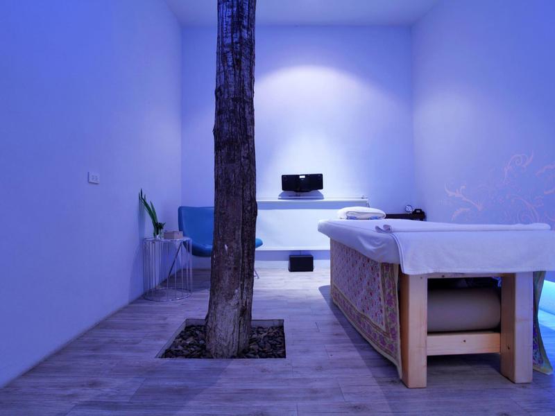 Minimalistischer Raum mit blauer Beleuchtung, Massageliege und Baumstamm in der Mitte.