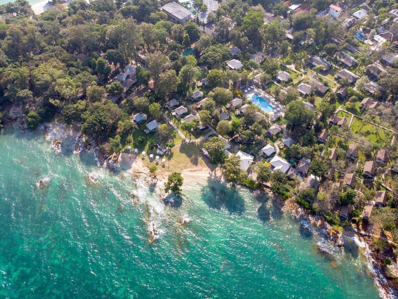 Luftaufnahme einer Küstenlinie mit Häusern, Bäumen und klarem, türkisfarbenem Wasser.