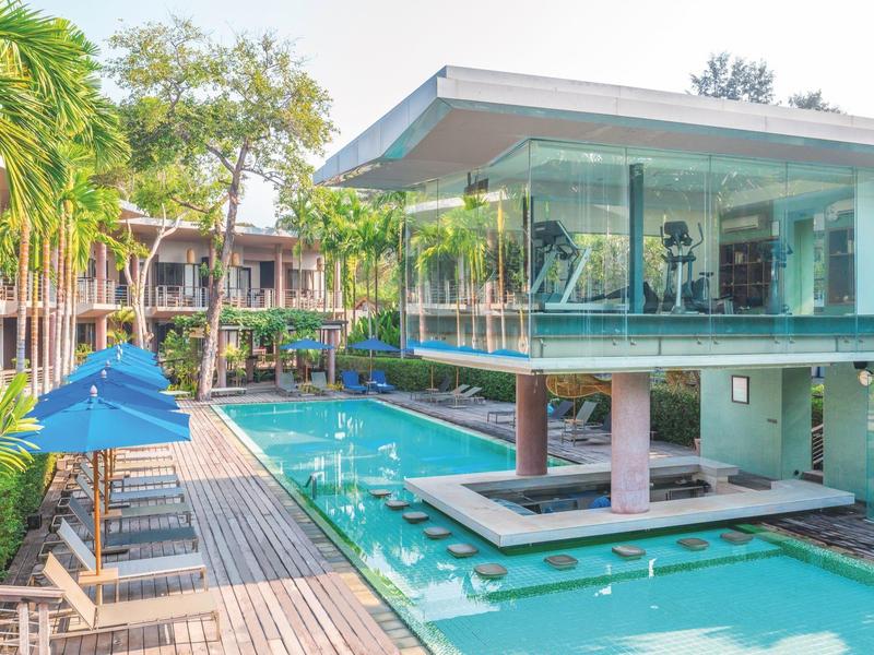 Moderner Poolbereich mit gläsernem Gebäude, Holzdeck, blauen Sonnenschirmen und grüner Vegetation.