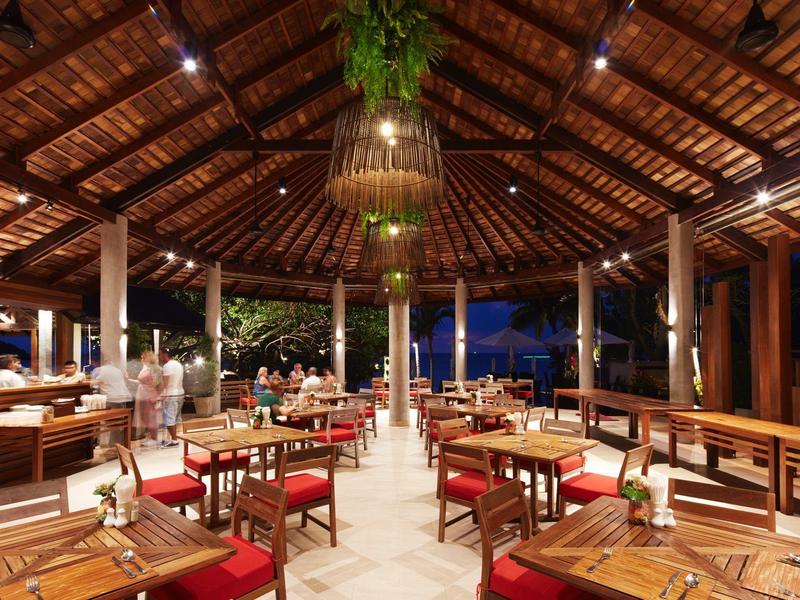 Offenes Restaurant mit Holzdach, Holztischen, roten Sitzkissen und warmen Lampen bei Abenddämmerung.