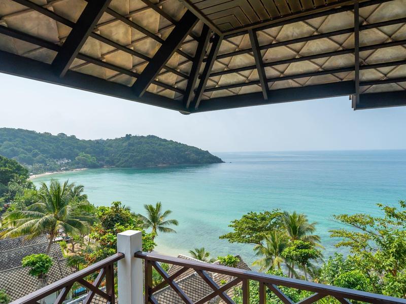 Blick von einer Terrasse mit Holzdach auf helle Sandstrände, türkisfarbenes Meer und bewaldete Hügel.
