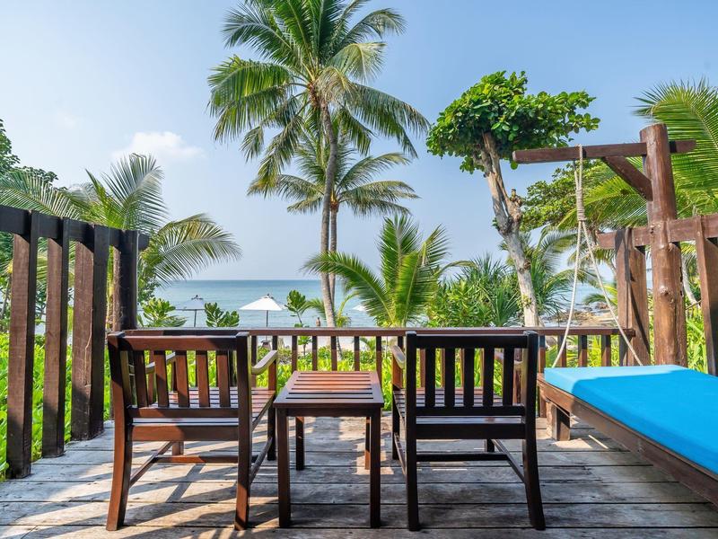 Holzterrasse mit Stühlen, Tisch und Liege, umgeben von Palmen mit Meerblick bei klarem Himmel.