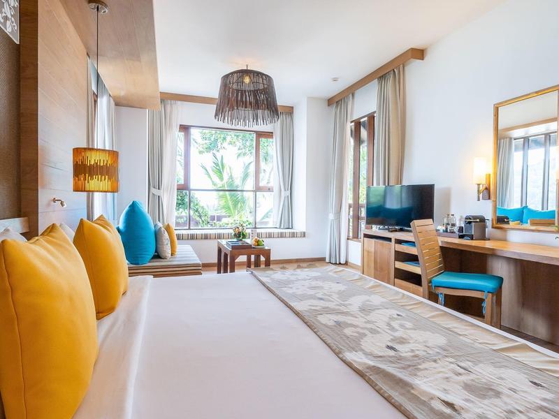 Helles Schlafzimmer mit großem Bett, gelben Kissen, Holzmöbeln und Fenster mit Blick ins Grüne.