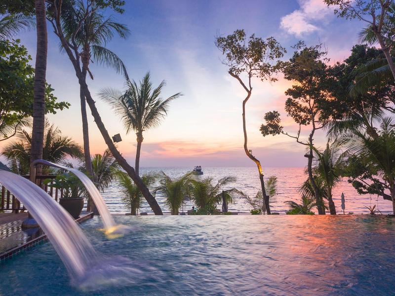 Infinity-Pool mit Wasserfall vor Palmen und Sonnenuntergang am Meer, ruhige tropische Atmosphäre.