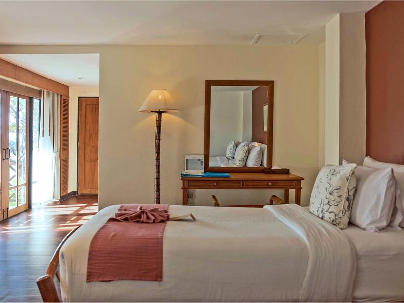 Helles Schlafzimmer mit Doppelbett, Spiegel, Stehlampe und großen Fenstern mit Blick ins Freie.