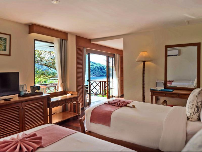 Hotelzimmer mit zwei Einzelbetten, Holzmöbeln, Fenster mit Blick auf See und Balkon, warme Beleuchtung.