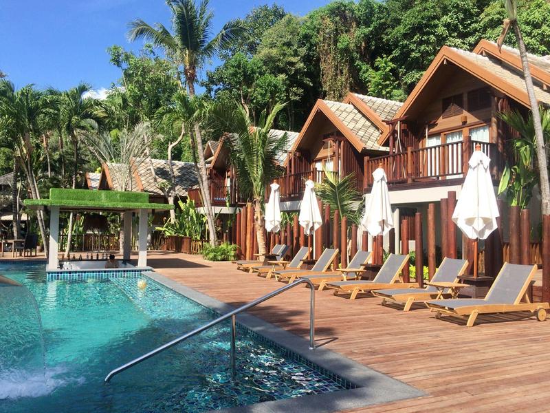 Hotel mit Holzbalkonen, Liegestühlen, weißen Schirmen und großem Pool vor grünen Palmen.