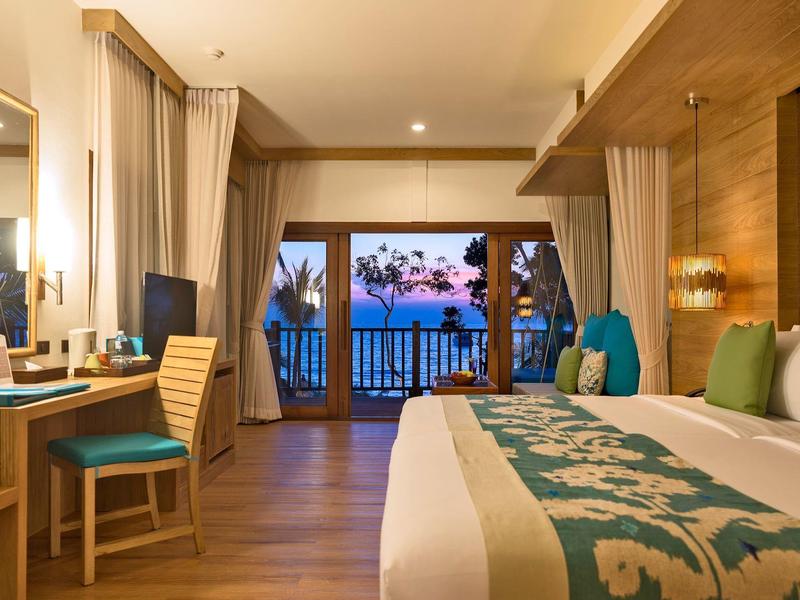 Helles Hotelzimmer mit großem Bett, Holzmöbeln, Balkon und Blick auf das Meer bei Sonnenuntergang.