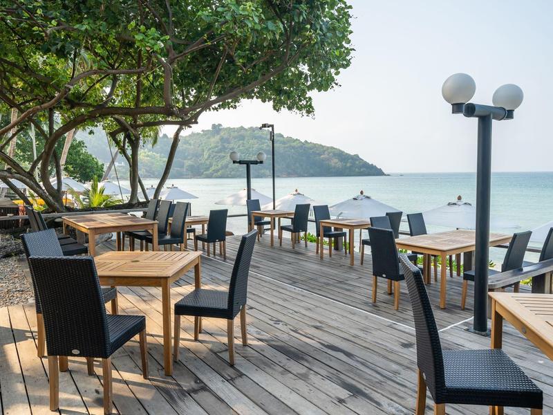 Draußenbereich mit Holzdeck, Tischen und Stühlen, Bäumen und Blick aufs Meer und Hügel im Hintergrund.