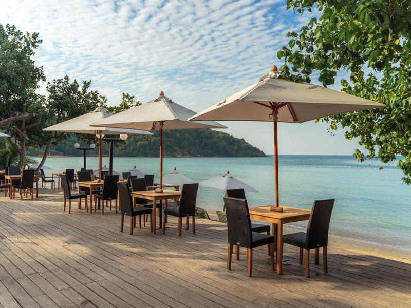 Holzterrasse am Meer mit Tischen, Stühlen und Sonnenschirmen unter blauem Himmel.